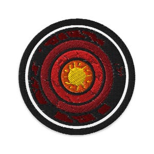 May include: Embroidered patch featuring a circular design with concentric rings of red and black. The center has a yellow sun-like symbol. The patch is bordered by a white stripe and a black outer edge. The design is detailed and textured.