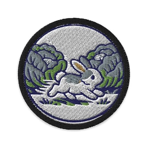 May include: Embroidered patch featuring a white rabbit running through a stylized landscape with gray and green elements. The design is circular, with a black border and a textured, off-white background.