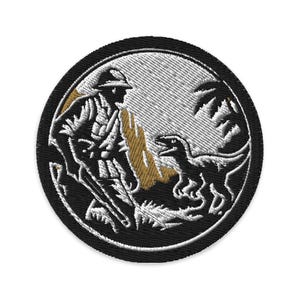 Op de afbeelding: Ronde geborduurde patch met een zwarte rand, die een soldaat en een dinosaurus in een junglescène afbeeldt. Het ontwerp maakt gebruik van witte en bruine steken om een gedetailleerd beeld van de figuren en de omgeving te creëren.