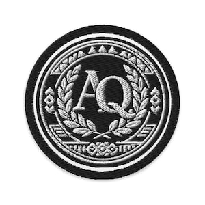 May include: A circular black embroidered patch with a white border. The patch features the letters "AQ" in a serif font, surrounded by a laurel wreath and decorative elements. The design is detailed and symmetrical.