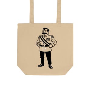 May include: Beige tote bag featuring a black and white illustration of a stern-looking man in a military uniform. The illustration includes a large mustache, medals, and a sash. The bag has two handles.