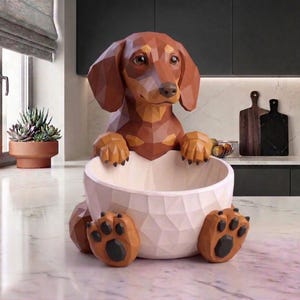 Op de afbeelding: Een bruine en tan teckel hond sculptuur met een geometrisch ontwerp, met een witte kom. De poten en kop van de hond zijn gedetailleerd en de kom heeft een getextureerd oppervlak. De sculptuur is een decoratief item voor in huis.