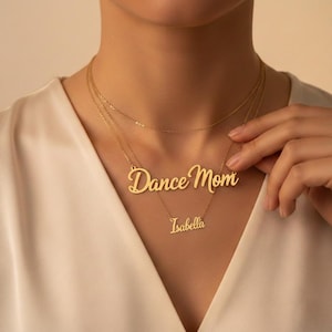 May include: Gold-colored layered necklaces. The top necklace is a delicate chain. The middle necklace says "Dance Mom" in cursive. The bottom necklace says "Isabella" in cursive. The necklaces are worn by a person wearing a white top.