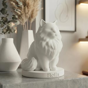 Puede incluir: Figura de gato de cerámica blanca con detalles de pelaje esponjoso, sentado sobre una base redonda. El nombre "Bella" está inscrito en la base. Un artículo decorativo para el hogar.