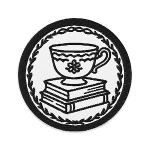 May include: Embroidered patch featuring a teacup with a floral design, resting on a stack of books. The design is in black and white, surrounded by a black border with a leaf and chain pattern. The patch is circular.