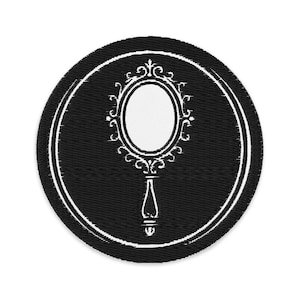 Puede incluir: Un parche circular negro con un diseño de espejo de mano blanco y ornamentado. El espejo tiene forma ovalada con un marco y un asa decorativos. Un borde blanco rodea la imagen del espejo sobre un fondo negro.