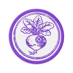 May include: A round, embroidered patch featuring a purple radish with leafy greens. The radish has a cross-shaped detail and is set against a white background, encircled by a purple border.