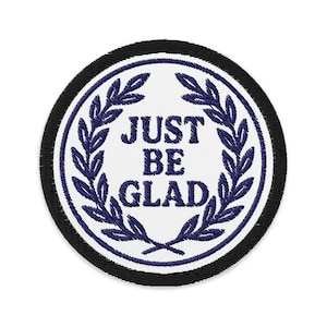 May include: Circular embroidered patch featuring the text "JUST BE GLAD" in navy blue, encircled by a navy laurel wreath on a white backdrop. The patch is edged with a black border.