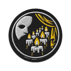 Op de afbeelding: Geborduurde patch met een wit masker links en brandende kaarsen rechts, tegen een zwarte achtergrond. De kaarsen hebben gele vlammen en witte was. Een gele gordijn is op de achtergrond. De patch is rond met een witte rand.
