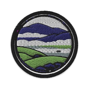 Op de afbeelding: Geborduurde patch met een zwarte cirkelvormige rand, die een landschap weergeeft. Het ontwerp toont blauwe bergen, witte wolken, groene heuvels en een klein zwart dierensilhouet, mogelijk een beer of wolf.