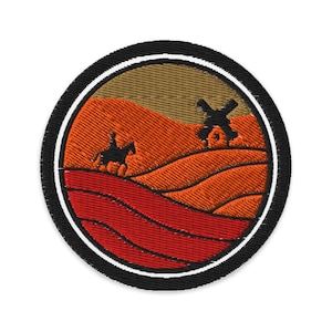 Op de afbeelding: Geborduurde ronde patch met een landschap met een ruiter te paard en een windmolen. Het ontwerp maakt gebruik van oranje, rode en bruine tinten, met een zwarte rand en een witte binnenring. Ideaal voor kleding of accessoires.