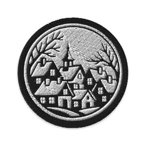May include: Embroidered patch featuring a black and white village scene within a circular frame. The design includes stylized houses, trees, and a textured background, suitable for jackets, bags, or other fabric items.