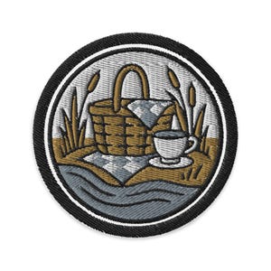 May include: Embroidered patch featuring a picnic scene. A brown wicker basket with a handle sits on a checkered blanket, next to a teacup and saucer. Reeds and water are also depicted, all within a circular frame.