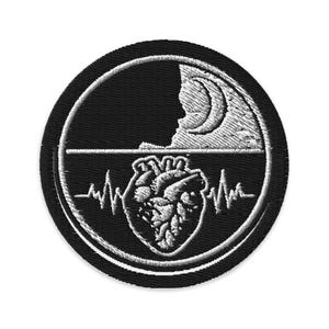 Op de afbeelding: Zwart en wit geborduurde patch met een cirkelvormig ontwerp. De bovenste helft toont een maan en een silhouet. De onderste helft toont een hart met een hartslaglijn. De patch is rond.