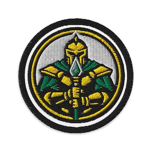 May include: Round embroidered patch with a warrior design. The warrior is depicted in gold and green armour, holding a spear. The design is set within a white and yellow circle, with a black border. Suitable for clothing.