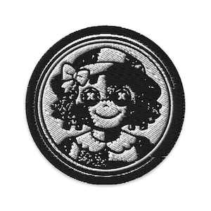 Może przedstawiać: Okrągła, haftowana naszywka w kolorze czarno-białym, przedstawiająca stylizowaną twarz lalki z oczami w kształcie krzyża, kokardą i uśmiechniętymi ustami. Wzór zamknięty w białym okręgu i czarnej obwódce. Odpowiednia do odzieży.