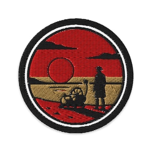 Op de afbeelding: Geborduurde patch met een woestijnlandschap met een rode zon, een figuur en een voertuig met wielen. De patch heeft een zwarte rand met een witte binnenrand. Het ontwerp is cirkelvormig.