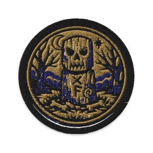 Könnte beinhalten: Gestickter Aufnäher mit einer goldfarbenen, schädelartigen Figur und einem Kreuzdesign, in einem goldenen Kreis. Das Design umfasst einen Mond, Sterne und blaue Akzente auf schwarzem Hintergrund.