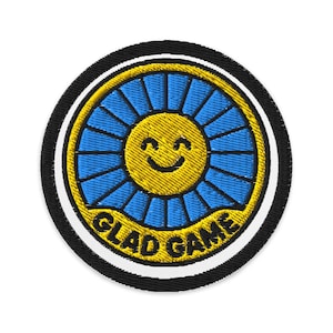 May include: Embroidered patch featuring a smiling yellow sun with blue rays, encircled by a gold border and a black and white outer ring. The text "GLAD GAME" is arched below the sun.