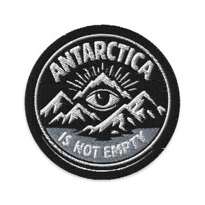 May include: Embroidered patch with the words "ANTARCTICA" and "IS NOT EMPTY" in white thread on a black circular background. The design includes a mountain range with an eye in the center, and sun rays.