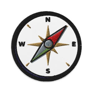 May include: Embroidered patch featuring a compass design. The compass rose is gold with red and green accents, set against a white background. The cardinal directions N, E, S, and W are clearly marked. The patch has a black border.