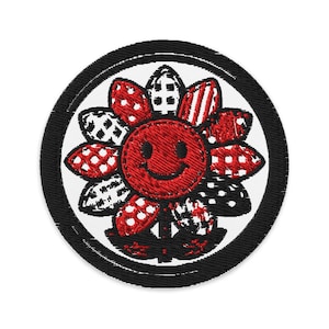 Może przedstawiać: Okrągła, haftowana naszywka z uśmiechniętym czerwonym kwiatem i czarną obwódką. Płatki kwiatu mają wzory w kolorze czerwonym, białym i czarnym, w tym grochy, paski i szachownice. Naszywka ma białe tło.