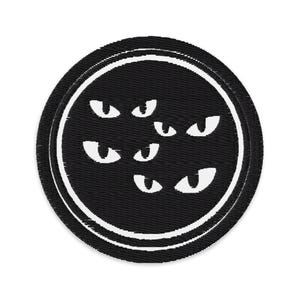 May include: A circular black patch with a white border. The patch has a design of several white, stylised eyes inside a black circle. The eyes are of different sizes and orientations, creating a spooky or whimsical effect. The patch is likely for clothing or accessories.