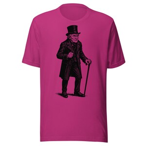 May include: A magenta t-shirt featuring a black illustration of a stern-looking man in a top hat, coat, and holding a cane. The graphic is a detailed, vintage-style drawing. The shirt is a solid color, with a crew neck and short sleeves.
