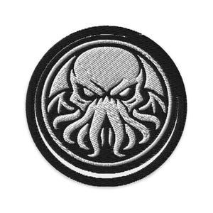 May include: Embroidered patch featuring a stylized, white Cthulhu design on a black circular background. The design includes a skull-like face with tentacles and wings, surrounded by a double border. The patch is suitable for jackets, bags, or other fabric items.
