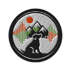 Op de afbeelding: Geborduurde patch met een zwart silhouet van een hond voor een bergketen. Het ontwerp bevat een oranje zon, groene en oranje verticale lijnen en een witte achtergrond, alles binnen een zwarte cirkel.