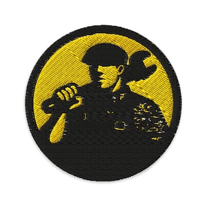 May include: Embroidered patch featuring a mechanic holding a wrench, set against a yellow circular background. The mechanic is depicted in black, with details of the uniform visible. The patch has a black border.