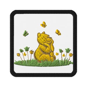 May include: Embroidered patch showing a yellow bear looking upwards, with yellow and green butterflies and flowers. The patch has a white background and a black border, creating a charming, whimsical design.