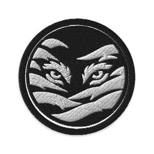 May include: Embroidered patch featuring a stylized design of a tiger's eyes. The patch is circular, with a black background and white thread used for the detailed eye and stripe design. The patch has a black border.
