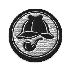 Könnte beinhalten: Gestickter Aufnäher mit einer schwarzen Silhouette eines Detektivhuts und einer Pfeife in einem weißen Kreis auf schwarzem kreisförmigen Hintergrund. Das Design hat ein strukturiertes, genähtes Aussehen, geeignet für Kleidung oder Accessoires.
