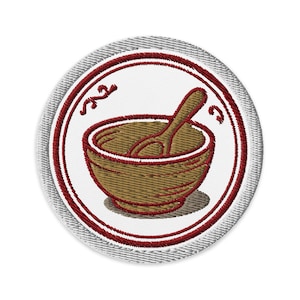 Può includere: Toppa ricamata raffigurante una ciotola marrone con un cucchiaio, su sfondo bianco. La ciotola e il cucchiaio sono delineati in marrone, con un bordo rosso ed elementi decorativi. La toppa ha un bordo grigio strutturato.