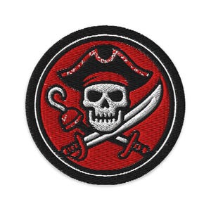 May include: Embroidered patch with a pirate skull wearing a tricorn hat, crossed swords, and a hook. The design is set within a red circle, with a black border. A classic pirate theme.