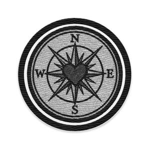 May include: A round, embroidered patch featuring a compass rose design with a heart at the center. The compass points are labeled N, E, S, and W. The patch has a black and white color scheme with a textured background.
