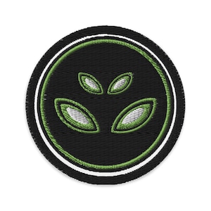May include: Embroidered patch featuring a stylized alien face with three green oval eyes on a black background. The design is encircled by a white and green border, creating a circular shape. The patch is likely for clothing or accessories.