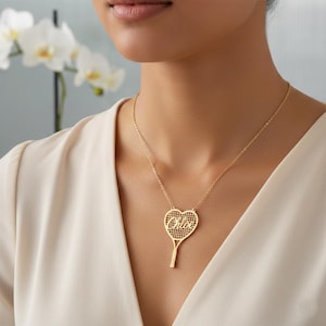 Puede incluir: Un collar con colgante de raqueta de tenis dorado con una cabeza en forma de corazón y el nombre "Chloe" inscrito. El collar se lleva alrededor del cuello y cuelga de una delicada cadena. El fondo está borroso, con un toque de flores blancas.