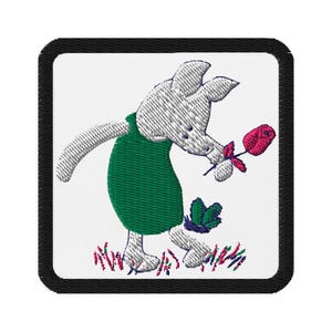 Op de afbeelding: Geborduurde patch met een cartoonkonijn in een groene jurk, met een rode roos in de hand. Een groene vlinder en kleurrijk gras zijn ook op de patch geborduurd. De patch heeft een zwarte rand en een witte achtergrond.