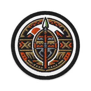 May include: Embroidered patch with a circular design featuring a central shield and spear motif. The patch has a black border, a white inner border, and intricate details in orange, brown, and black.