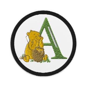 May include: Embroidered patch featuring a yellow bear holding a honey pot next to a green letter "A". The patch is circular with a white background and a black border. The design is a cartoon style.