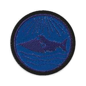 May include: Embroidered patch featuring a stylized purple fish above blue waves, set within a blue circle. The design includes radiating lines and a black border, creating a nautical or aquatic theme.