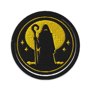 Könnte beinhalten: Runder, gestickter Aufnäher mit einer schwarzen Silhouette einer gewandeten Figur mit Stab, vor einem gelben Mond. Das Design umfasst gelbe Sterne und einen kreisförmigen Rand, alles auf schwarzem Hintergrund.