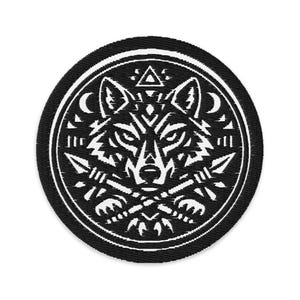 May include: A circular, black embroidered patch with a white wolf design. The wolf's head is central, encircled by a border with arrows, crescent moons, and geometric patterns. The patch has a textured finish.