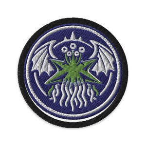Op de afbeelding: Geborduurde patch met een groen, stervormig wezen met meerdere ogen en tentakels, tegen een blauwe achtergrond. Het ontwerp bevat witte vleugels en is omlijst door een zwarte rand. Ideaal voor jassen, tassen of andere stoffen items.