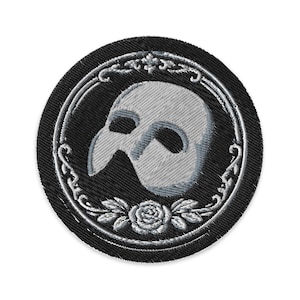 Op de afbeelding: Geborduurde patch met een grijze Phantom of the Opera-masker in een decoratief zilveren frame op een zwarte cirkelvormige achtergrond. Het ontwerp bevat een roos en bladeren aan de onderkant. Ideaal voor jassen, tassen of andere stoffen items.