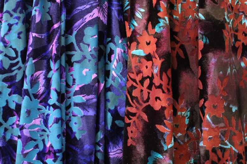 May include: Close-up of three fabric swatches with floral patterns. The fabric is a dark blue velvet with a teal and purple floral design, a dark purple velvet with a teal and purple floral design, and a dark red velvet with a teal and red floral design.