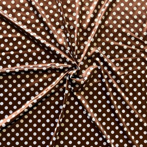 May include: Brown fabric with a repeating pattern of white polka dots. The fabric is folded and draped, creating shadows and highlights. The material has a slight sheen, suggesting a satin or silk-like texture. Ideal for sewing projects.