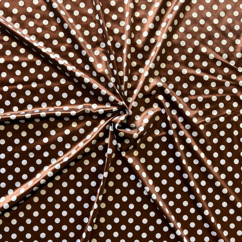 May include: Brown fabric with a repeating pattern of white polka dots. The fabric is folded and draped, creating shadows and highlights. The material has a slight sheen, suggesting a satin or silk-like texture. Ideal for sewing projects.